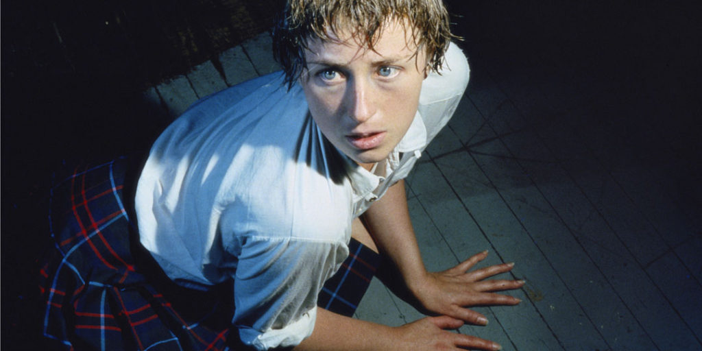 A photograph of the artist Cindy Sherman dressed in an outfit resembling a Catholic school uniform, with a white button down shirt and navy plaid shirt. Her short hair is sweaty or wet and she squats on her hands and knees in anticipation, looking beyond the frame with an expression of fear or dread.