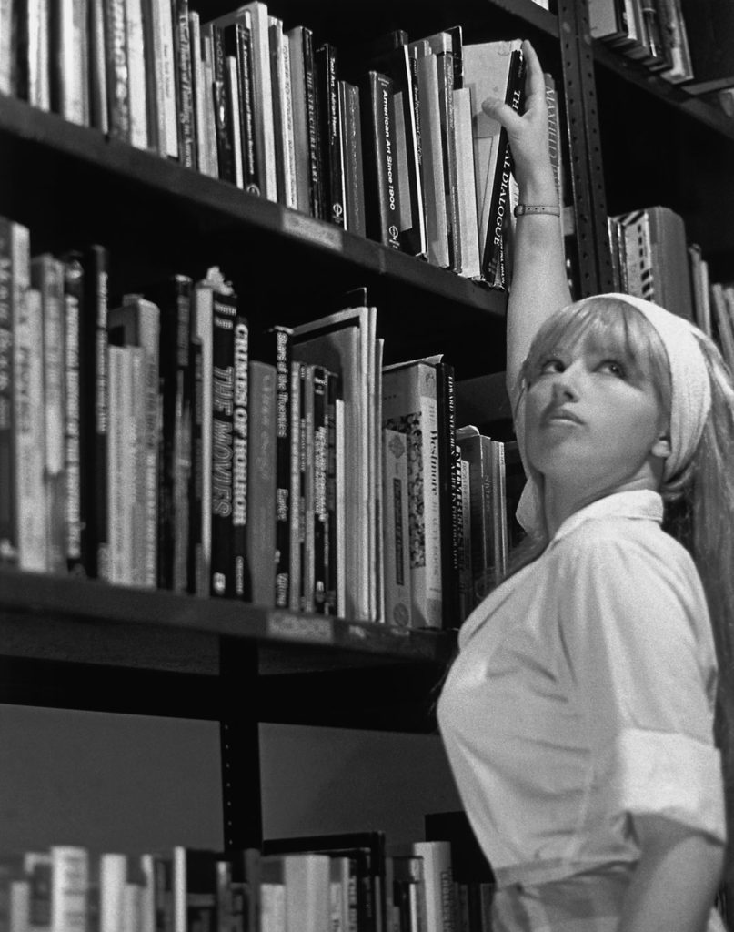A woman resembling Brigitte Bardot, with long bangs and a scarf, reaches up to lift a book off a shelf.