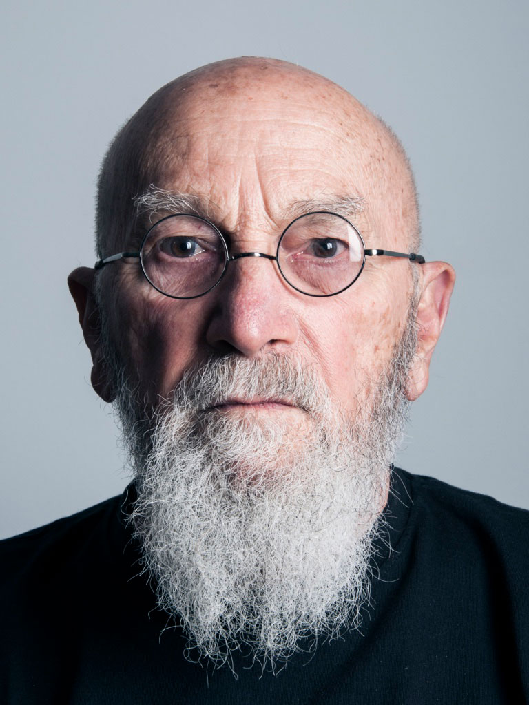A photograph of the artist Rafael Ferrer. He wears a dark shirt and round glasses and has a medium-length gray beard.
