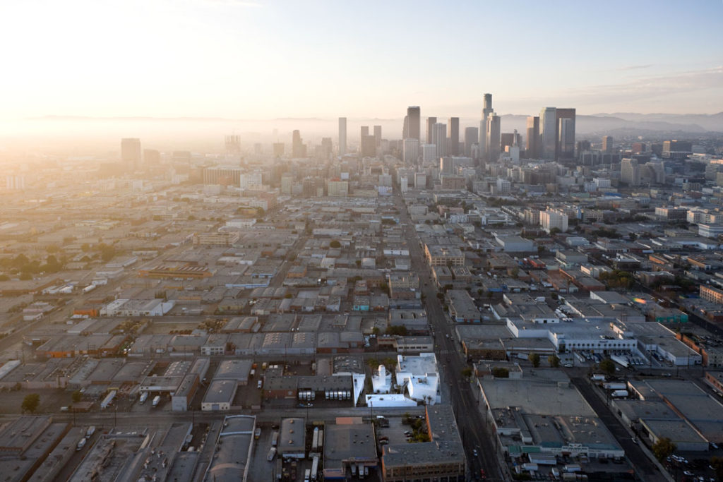 A birds eye view of the Inner-City Arts building. Located in the heart of Skid Row, the campus is a series of bright white buildings surrounded by uniform grey.  In the near distance is the beautiful outline of the Downtown Los Angeles skyline. 