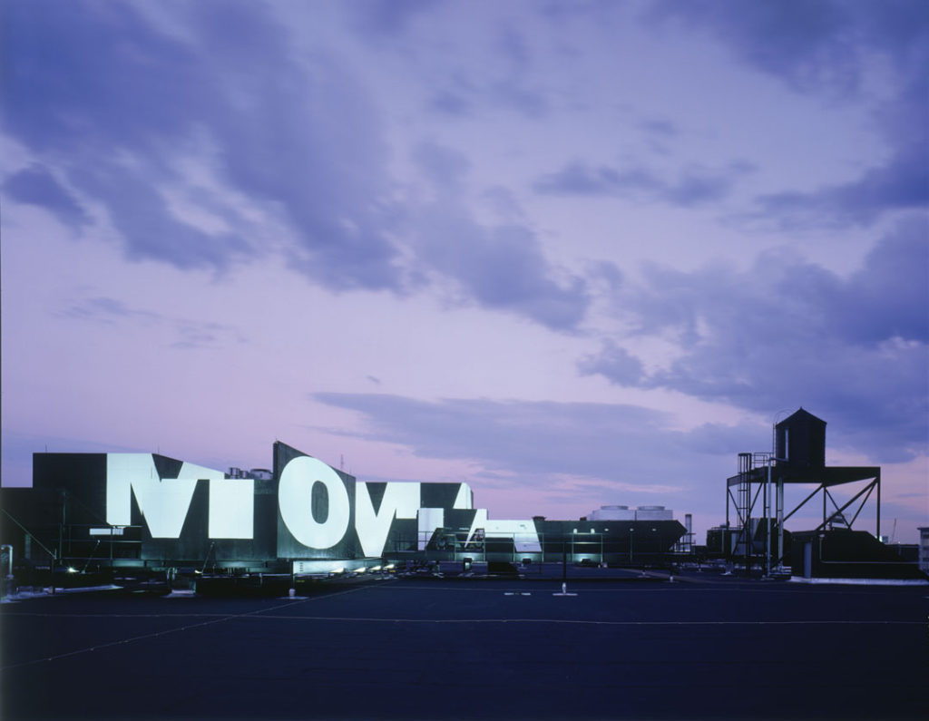 A dramatic evening shot of MoMA QNS.  We see the silhouette of the buildings which were originally the Swingline Staples factory, made up of a series of black boxes.  The MoMA logo appears in bright, bold white across the different shapes, similar to how projected light would appear.  To the right of the building we see the silhouette of a historic water tower.