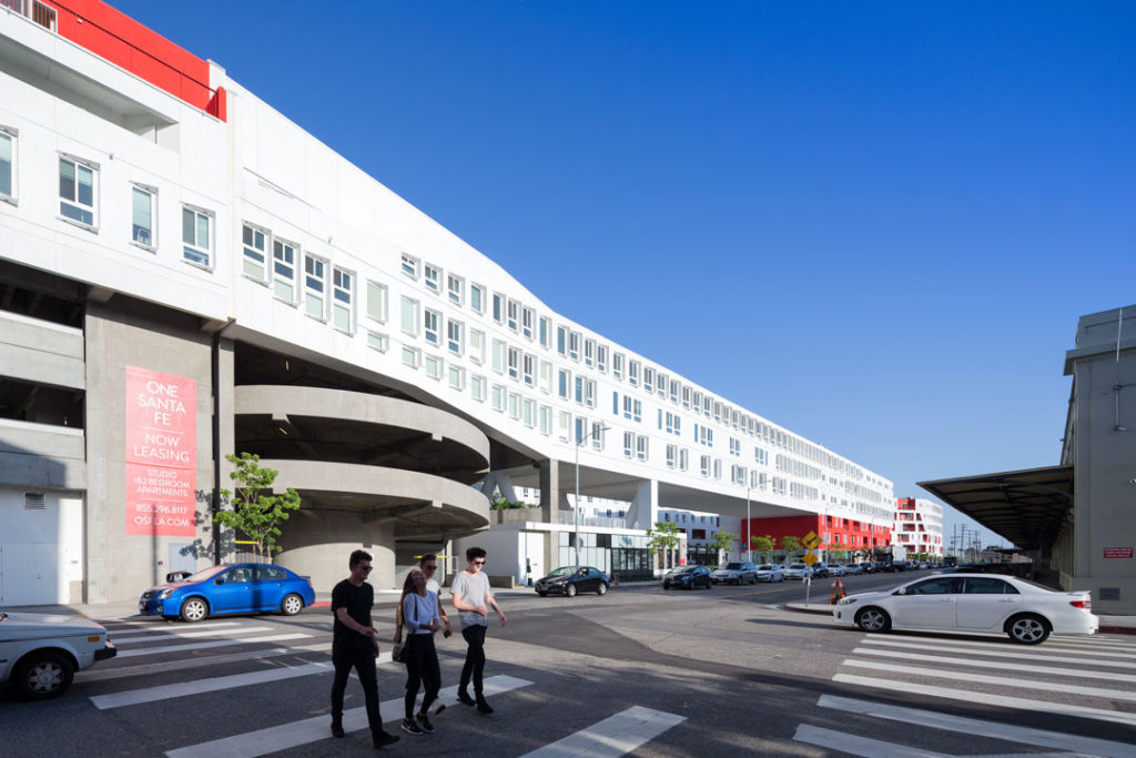 An exterior view of One Santa Fe, designed by Michael Maltzan. The building is made up of a series of long, white buildings with red accents that sit above the street. This view, shot from across the street, demonstrates how the buildings stretch far down the block and contain interesting nooks of space, including balconies and an open courtyard that is connected to retail spaces at street level.