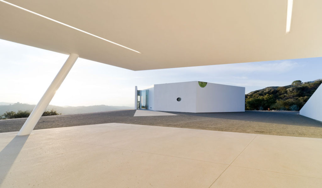 A photograph of an exterior living space at the Pittman Dowell residence.  From this view, we stand on a covered concrete pad facing a white, angular building and natural hills beyond.  In the distance is a ridge of mountains.  The image is punctuated by strong diagonal angles, drawing our view toward the horizon.