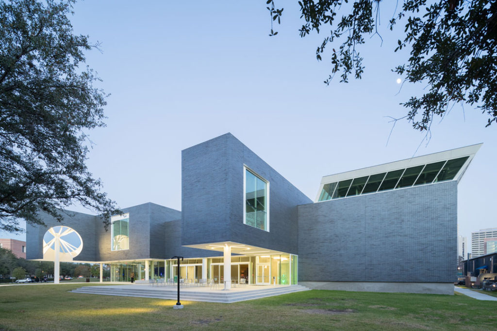 An exterior photograph of Rice University Moody Center for the Arts, designed by Michael Maltzan. The brick building is made up of a playful, quadrangle design.  The first floor includes a large pedestrian plaza with shaded areas from the cantilevered second-story areas that extend over it.  The entrance is made up of floor-to-ceiling glass, giving the space lightness and openness.  Large picture windows punctuate the façade.