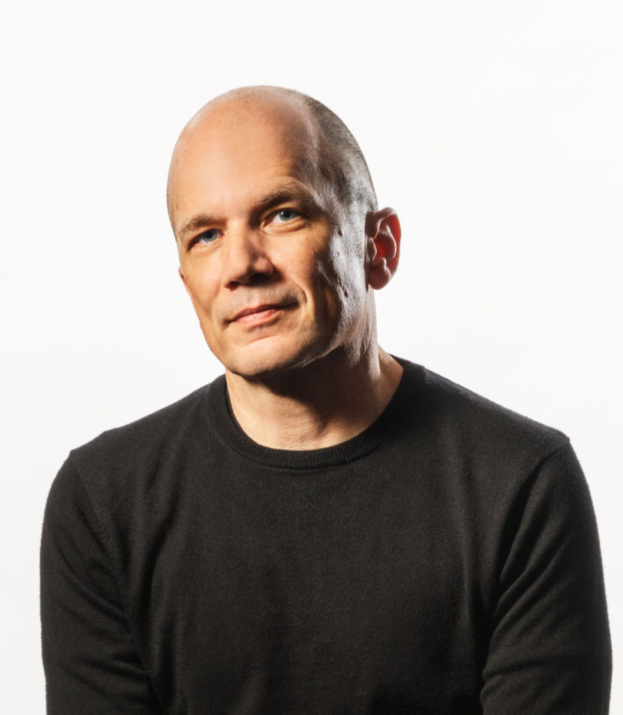 A headshot of architect Michael Maltzan. He is a white man with blue eyes and a shaved head. He wears a plain black sweatshirt and smiles in a quiet, friendly way at the camera.