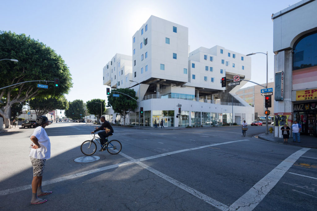 An exterior shot of the Star Apartments building, designed by Michael Maltzan. The white building has an interesting stacking design, with glass windows for the public health facilities at street level, an open-air plaza on the second level, and terraced floors of apartments above. 