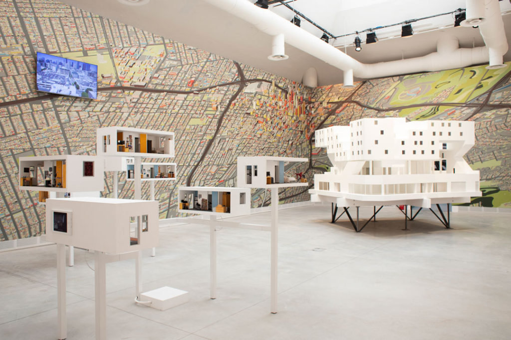 An installation photo of Michael Maltzan’s Star Apartments exhibition at the Venice Biennale. In the foreground are models of individual apartments. We’re not close enough to see the detail, but their interiors vary in content, color, and size. Further into the room is a model of the entire building, a striking white structure made from different stacking forms.  The walls are covered in an illustrated color map of Los Angeles.  A video monitor displays an image of the downtown skyline.