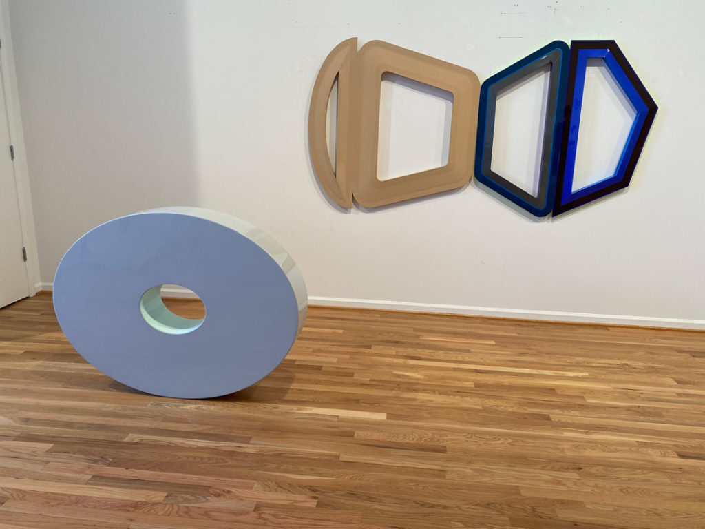 A photo of two pieces of Beverly Fishman’s artwork in her studio, taken. One of her polypharmacy compositions hangs on a white wall. It features four outlined shapes in shades of light brown and dark blue inspired by fragments of pharmaceuticals. One of Fishman’s sculptures rests on the hardwood floor nearby. It is a light blue oval with glossy edges and an aperture in the center.