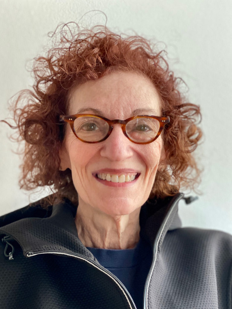 A headshot of artist Beverly Fishman taken by Matthew Biro. She is a white woman with curly reddish-brown chin-length hair. Her glasses match her hair. She is wearing a blue T-shirt with a gray zip-up athletic jacket over it.