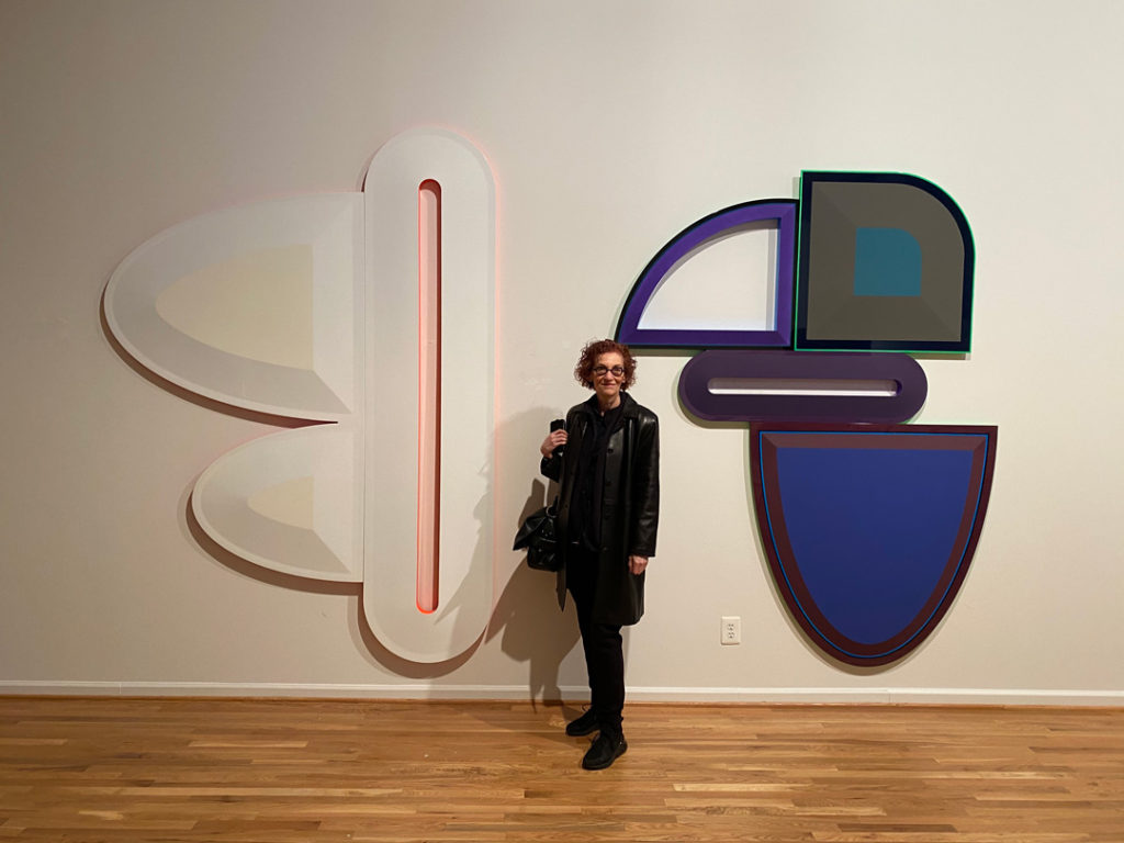 Photo of artist Beverly Fishman standing in front of her paintings Untitled (Chronic Pain, Pain, Pain) and Untitled (Pain, Opioid Addiction, Bipolar Disorder, Muscle Spasms), taken by Matthew Biro. The two large pieces are hanging on a white wall. Fishman is a white woman with reddish brown hair. She is wearing all black, including her jacket, shirt, pants, shoes, purse, and glasses. She is holding a cell phone.