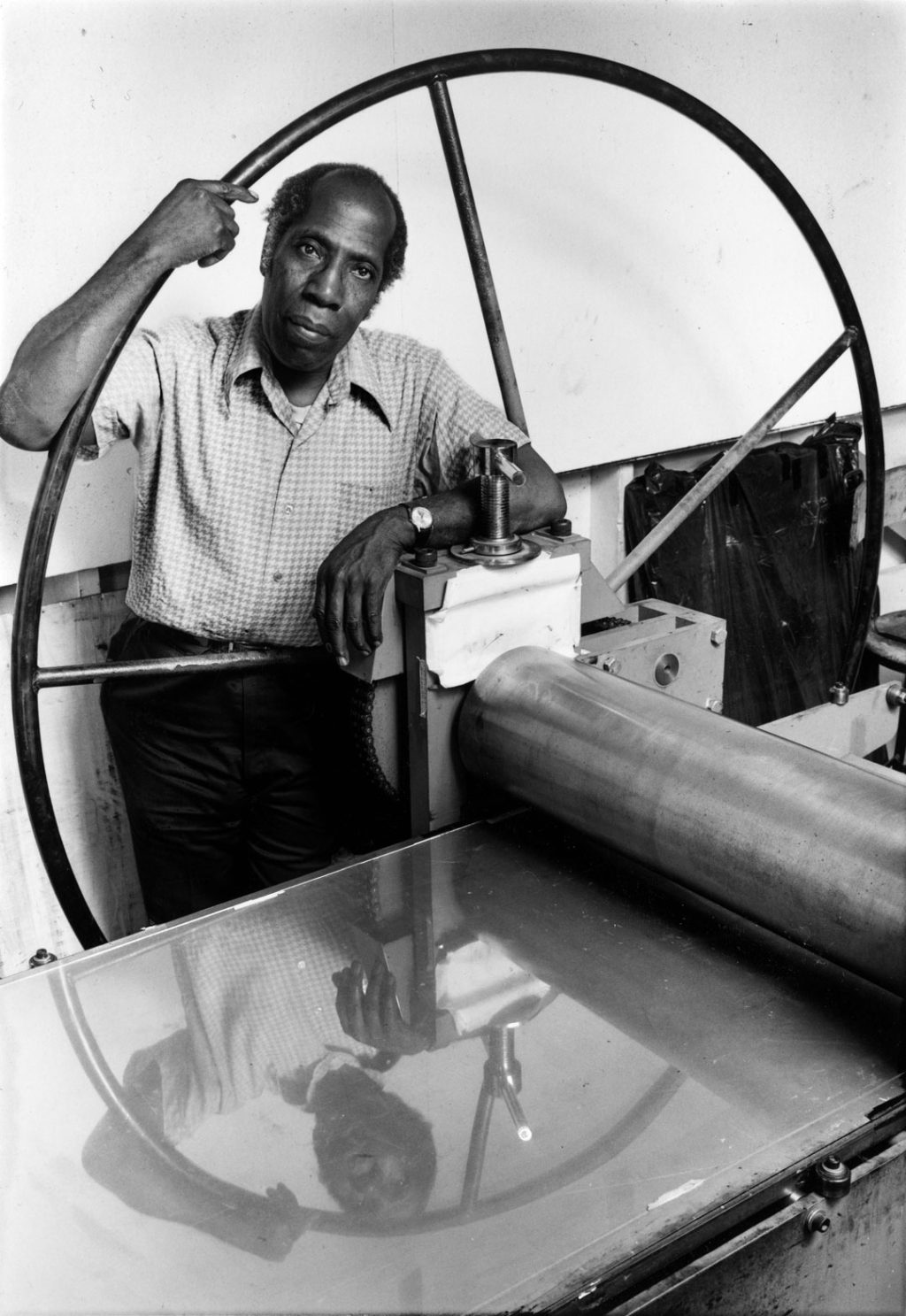 A photo of artist and printmaker Robert Blackburn taken by Peter Sumner Walton Bellamy. In the portrait, Blackburn is posing with printmaking equipment and looking straight at the camera with a calm expression. He is wearing a short-sleeve button-down shirt, dark pants, and a watch. His reflection is visible in the printing surface.