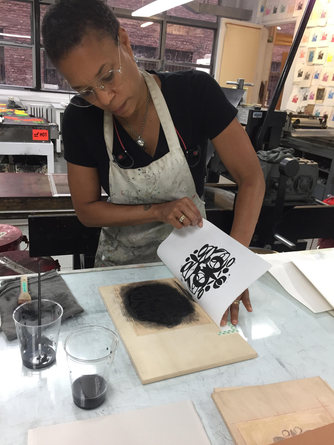 A photo of artist Karen J. Revis lifting a print to inspect it. She is a young Black woman with short hair, wearing wire-rimmed glasses, a black T-shirt, and a work apron. She is working in a room at the EFA Robert Blackburn Workshop that is full of different printing machines.
