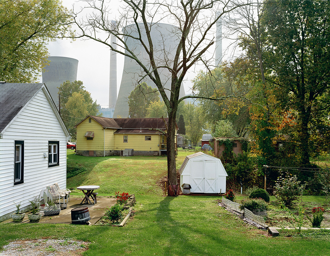 A color photo of two small houses, one white and one yellow, connected by a grassy yard with several trees. The white house has a small patio with several chairs, a table, and potted plants. A white shed is next to a tree. The setting would be bucolic, except for the towering concrete power plant stacks and chimneys just beyond the houses. The 2004 photo was taken by Mitch Epstein and is part of his American Power project, which captured how energy influences American lives.