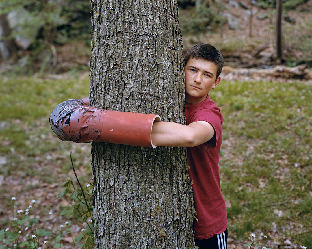 A color photo of Ashton Clatterbuck protesting the construction of a natural gas pipeline in Lancaster County, Pennsylvania. Clatterbuck is a young white man with dark hair. He is wearing a red t-shirt and hugging the trunk of a tree. His arms are inside a ‘sleeping dragon,’ a tool of civil disobedience made from PVC pipe and duct tape. He is looking straight at the camera with a serious expression on his face. The 2018 photo was taken by Mitch Epstein and is part of his Property Rights project.