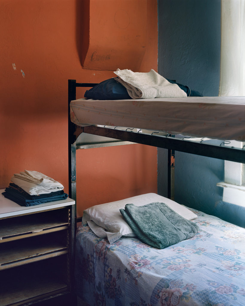 A color photo of a bunk bed in the corner of a room at Casa Vides, a halfway house for refugees in El Paso, Texas. The beds are neatly made with patterned sheets, and a pillow and folded towel is on each bed. A few folded sheets rest on a small shelving unit next to the bed. One wall of the room is painted cinnamon red and the other wall is a deep slate blue. The paint is chipped in a few places, but the light from a large window makes the room feel welcoming. This 2017 photo was taken by Mitch Epstein and is part of his Property Rights project.