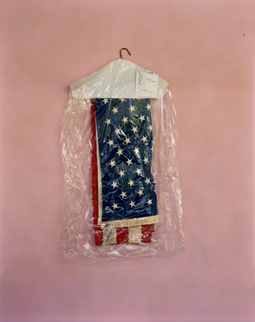 This color photo shows a folded American flag on a clothes hanger, hanging from a small nail on a pink wall. The flag is covered in a dry cleaner’s plastic bag, and a receipt attached to the hanger is marked “free.” The flag appears to be an antique; the stars and white stripes are yellowing with age. This photo was taken at Epstein Furniture in Holyoke, Massachusetts, a business that belonged to Mitch Epstein’s father. This 2000 photo was taken by Mitch Epstein and is part of his Family Business project.