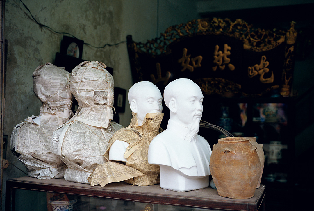 A color photo taken by Mitch Epstein in Hanoi, Vietnam, that shows four Ho Chi Minh busts displayed on a shelf next to two earthenware jugs. Two of the busts are completely wrapped in newspaper, one is partially wrapped, and one is unwrapped. This photo is part of Epstein’s Vietnam project, a photographic odyssey through modern-day Vietnam in the early 1990s.