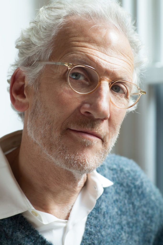 A headshot of photographer Mitch Epstein taken by Nina Subin. Epstein is a white man with light gray hair and a beard. He is wearing glasses and a white button-down shirt under a blue-gray wool V-neck sweater.