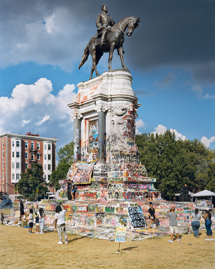 A color photo of the Robert E. Lee Monument in Richmond, Virginia. Above the monument, which depicts the Confederate general on his horse, there is a dark cloud in the sky. In 2020, following the Black Lives Matter protests, the statue was claimed by activists as a memorial for Black people who have died from police violence. The base of the monument is covered in colorful graffiti messages that say things like ‘Uplift black voices’ and ‘Tear it down.’ Signs posted on the monument or nearby read ‘BLM,’ ‘Say their names,’ and ‘Know Justice, Know Piece.’ A Black couple poses for a photo at the front of the monument, and off to one side, another woman poses for a photo. A few other Black men, women, and children look on. This 2020 photo was taken by Mitch Epstein and is part of his Property Rights project.