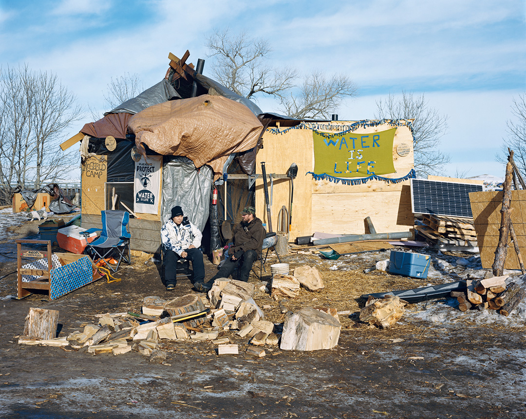 A color photo of two men in winter clothes sitting in camp chairs, smoking, in front of a structure made from wood and tarps at Standing Rock Sioux Reservation in North Dakota. Signs on the structure read ‘Rosebud Camp,’ ‘We Are Here to Protect’ and ‘Water Is Life.’ Tools, including shovels and an axe, hang on one side of the structure. Also nearby are coolers, chopped wood, a bucket, a camp chair, wooden pallets, and a solar panel. The 2017 photo was taken by Mitch Epstein and is part of his Property Rights project.