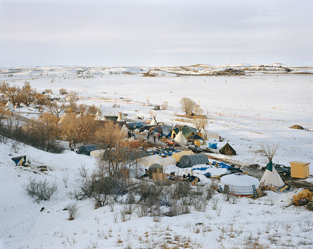 A color photo of a hilltop view of Sacred Stone Camp at Standing Rock Sioux Reservation in North Dakota. More than twenty structures make up the camp including tarp-covered longhouses, tipis, and a roundhouse. The trees are bare and the ground is covered in snow. Several people are visible, and a few cars are parked in the camp. An empty field and low, snow-covered hills are visible in the distance. This 2017 photo was taken by Mitch Epstein and is part of his Property Rights project.