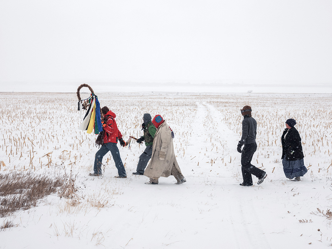 A color photo of marchers on a four-day prayer walk across the snowy plains of North Dakota in honor of the first anniversary of the closure of the primary encampment at Standing Rock, where indigenous people and activists protested the Dakota Access Pipeline. An Omaha Nation elder in a red jacket leads the march carrying a ceremonial eagle staff. Tire tracks lead into the distance. The 2018 photo was taken by Mitch Epstein and is part of his Property Rights project.